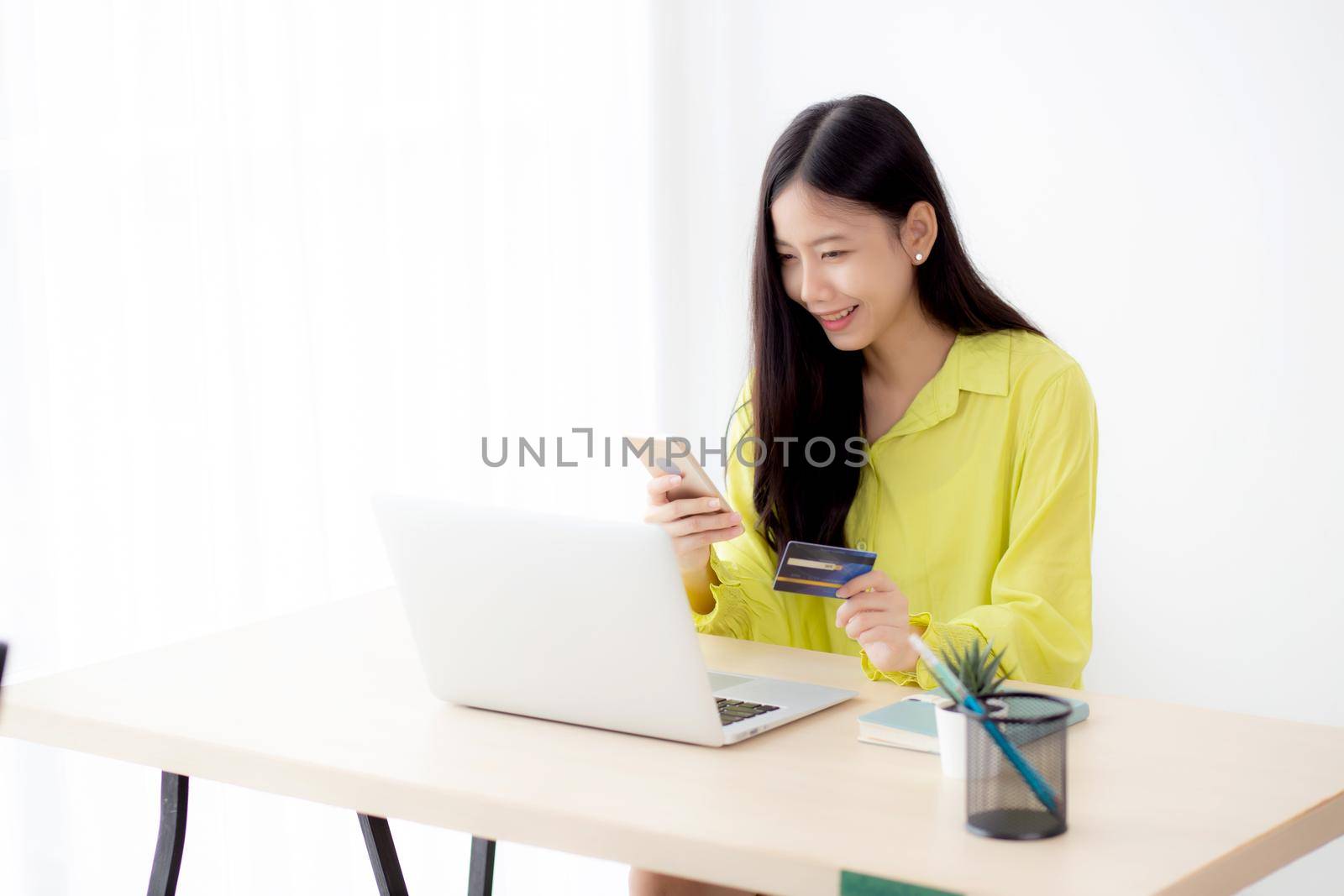 Young asian business woman using smart phone and holding credit card while online shopping and payment online with laptop computer on desk at home, female holding debit card, communication concept.