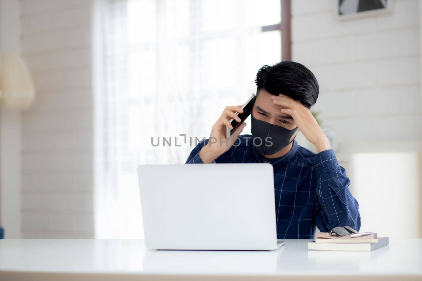 Young asian business man in face mask talking smartphone and work on laptop computer for protect covid-19, businessman quarantine work from home with notebook, social distancing and stay home.