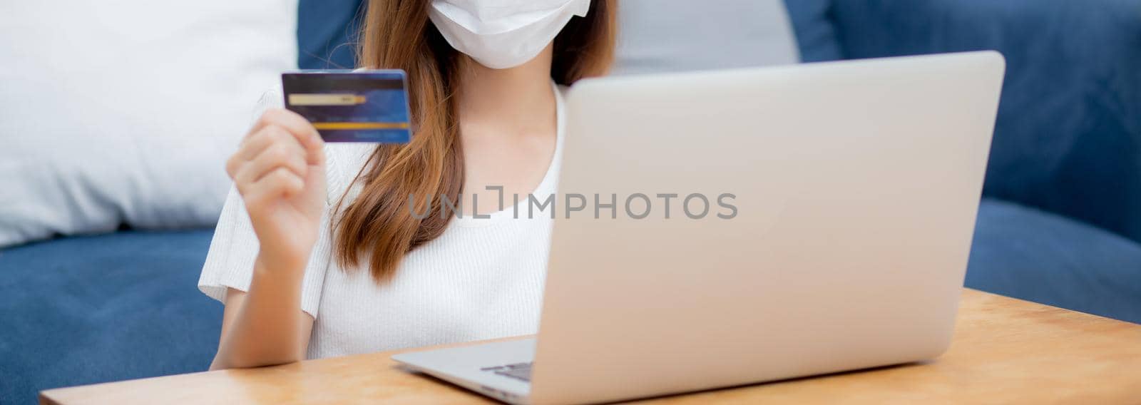 Young asian woman in face mask holding credit card shopping online with laptop computer buy and payment, girl using debit card purchase or transaction of finance, lifestyle and new normal concept. by nnudoo