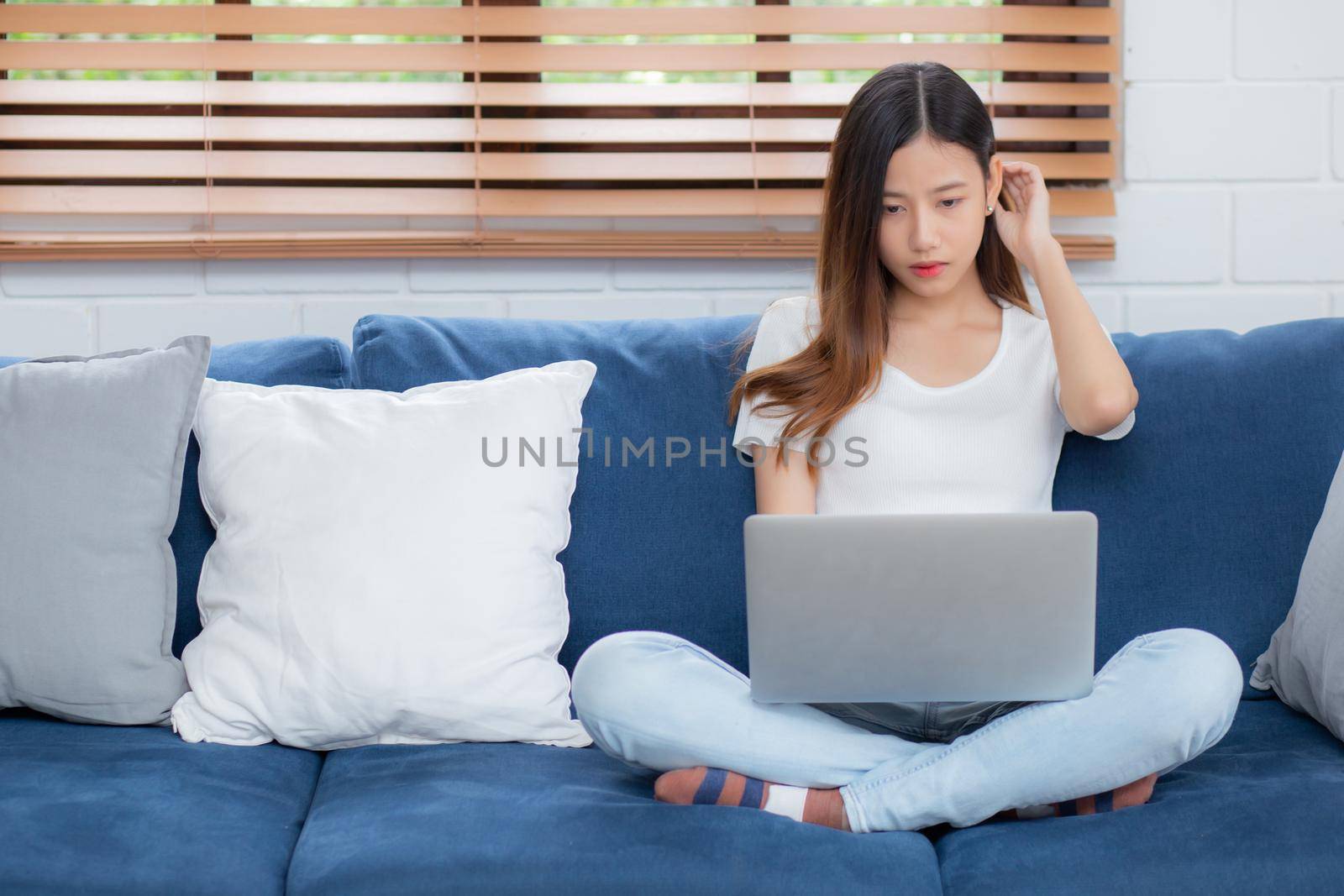 Young asian business woman smile and work from home with laptop computer online to internet on sofa in living room, freelance girl using notebook on couch with comfort, new normal, lifestyle concept.
