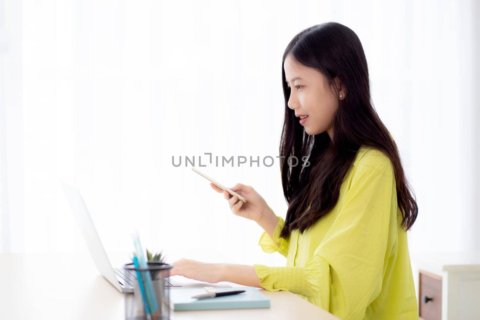 Young asian businesswoman working on laptop computer online on desk at home office, freelance woman looking message on smart phone and typing on notebook on table, business and communication concept.