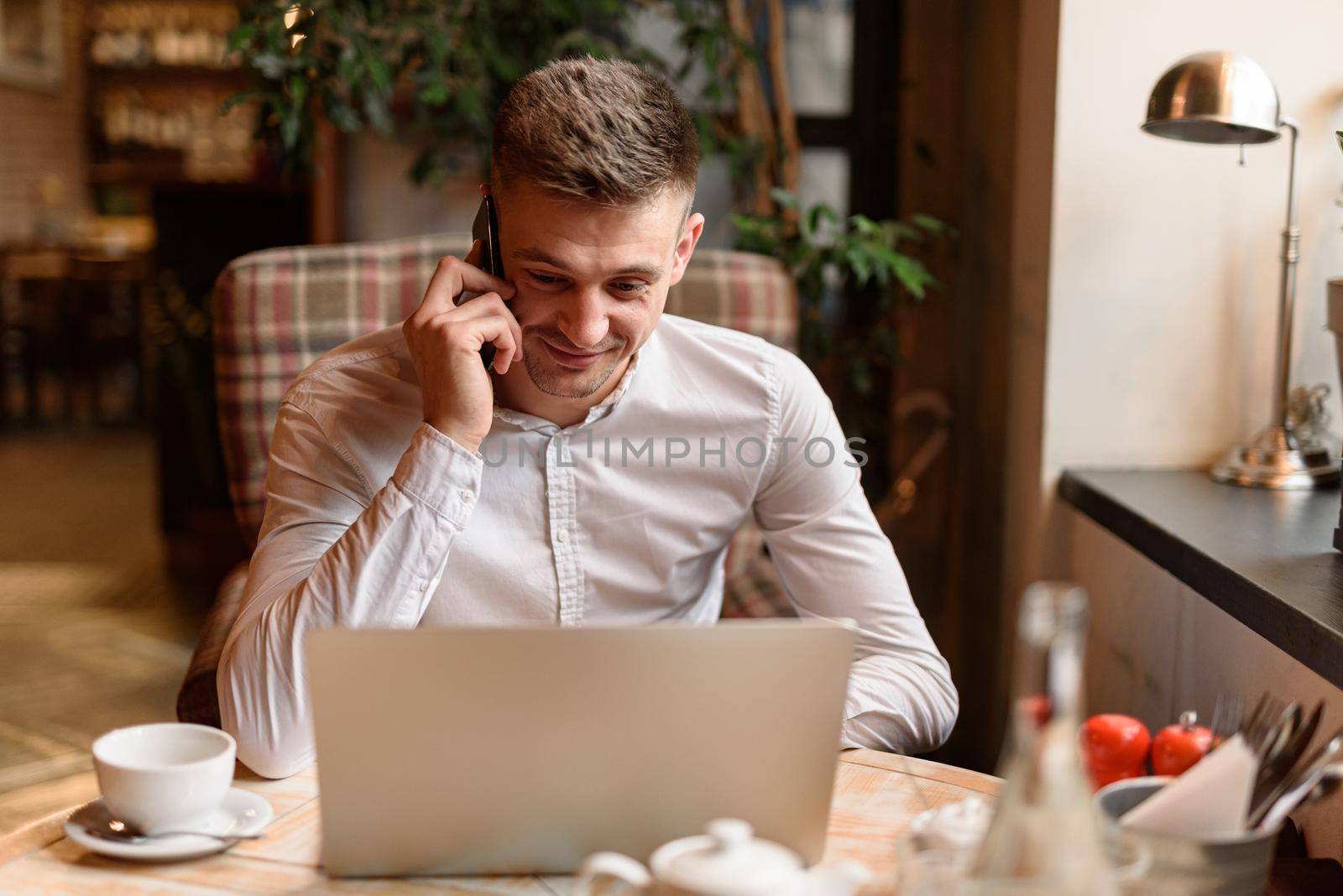 Waist up of smiling handsome businessman talking on smartphone while looking at laptop screen in cafe. Lifestyle concept