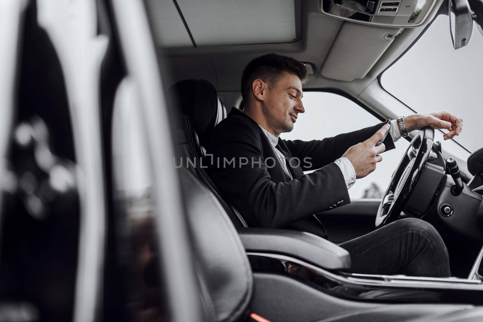 Side view of smiling handsome young man reading message on his smartphone while driving automobile. Rent and trade-in concept