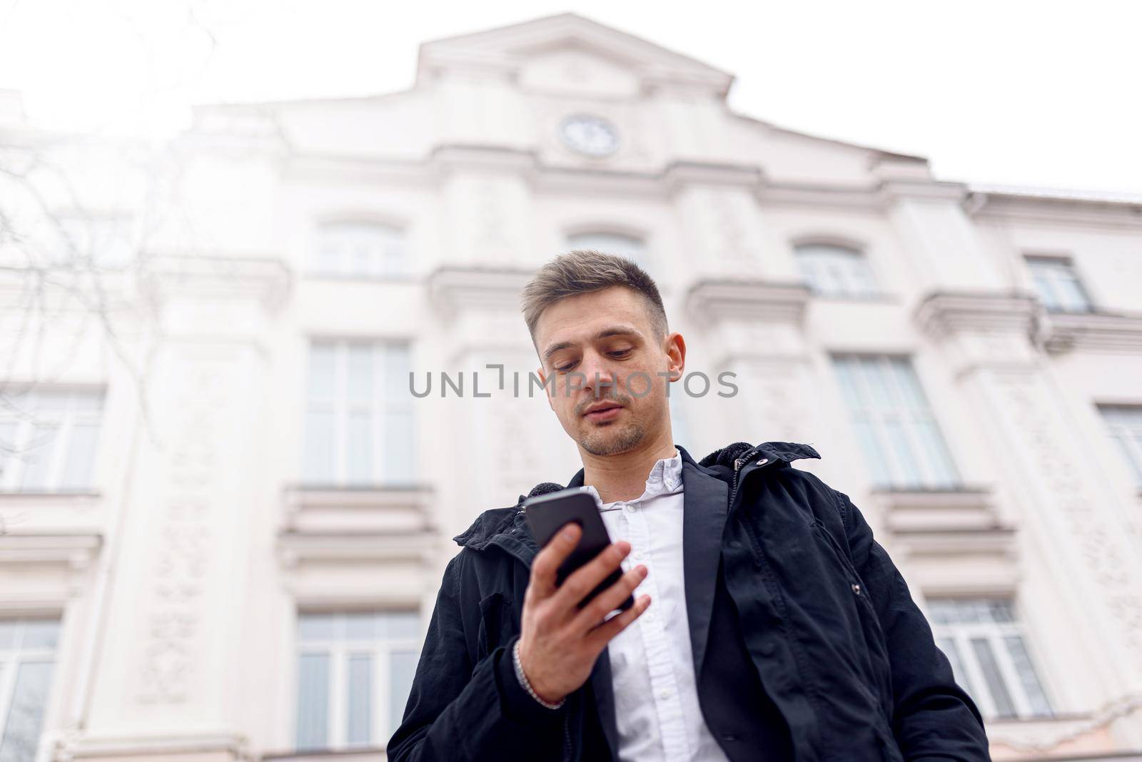 Waist up of handsome man in black jacket looking at smartphone screen in the city. Copy space. Lifestyle concept