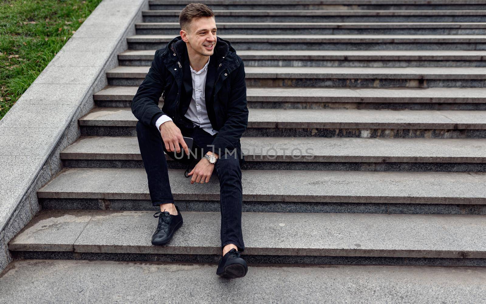 Top view of smiling young man with smartphone sitting on stairs in the city. Copy space. Lifestyle concept