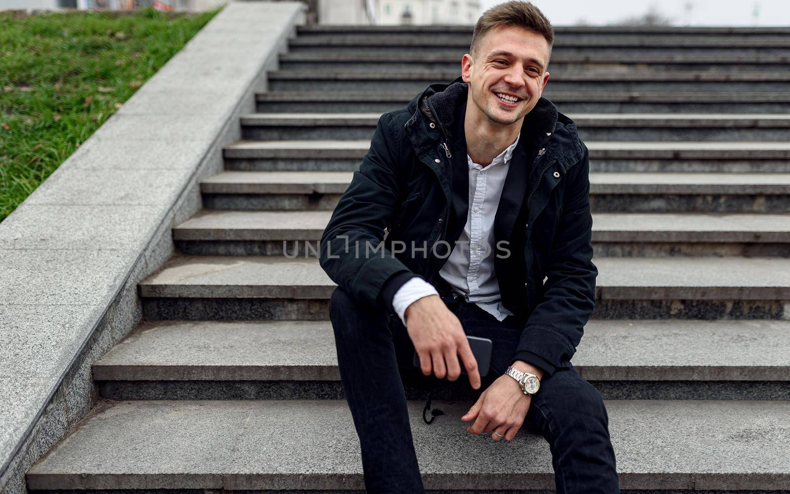 Waist up of happy handsome guy holding smartphone while sitting on stairs outdoors. Copy space. Lifestyle concept