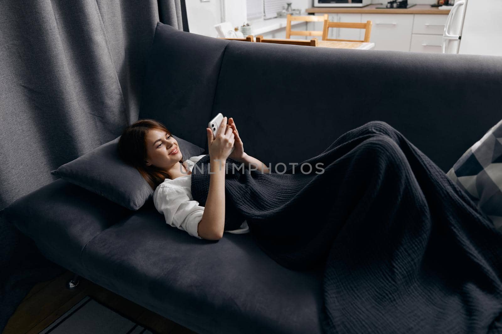a woman with a mobile phone lies on a gray sofa near the window in the background. High quality photo