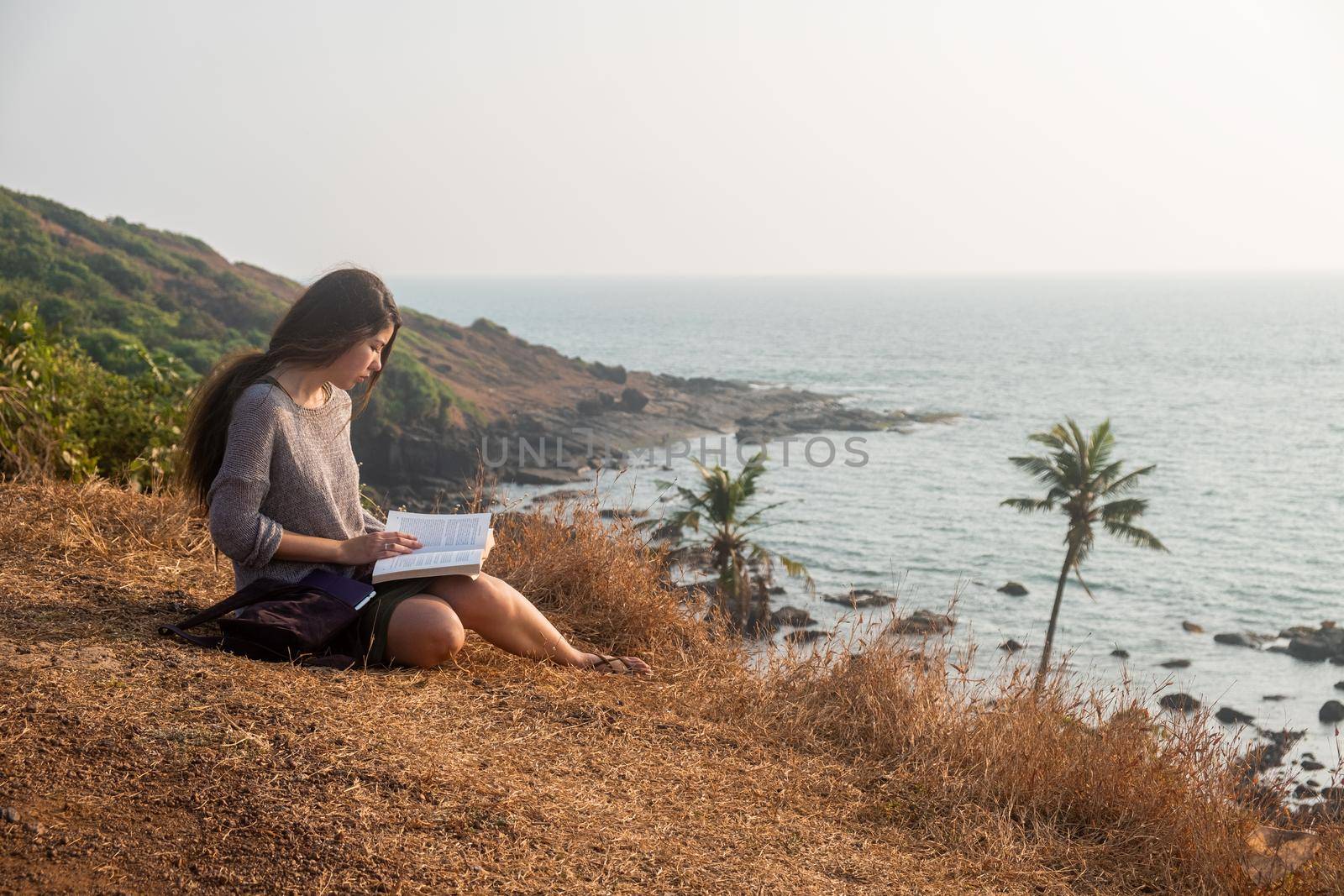 Attractive Asian woman is sitting on the edge of the mountain with a sea view, reading a book. Overall plan