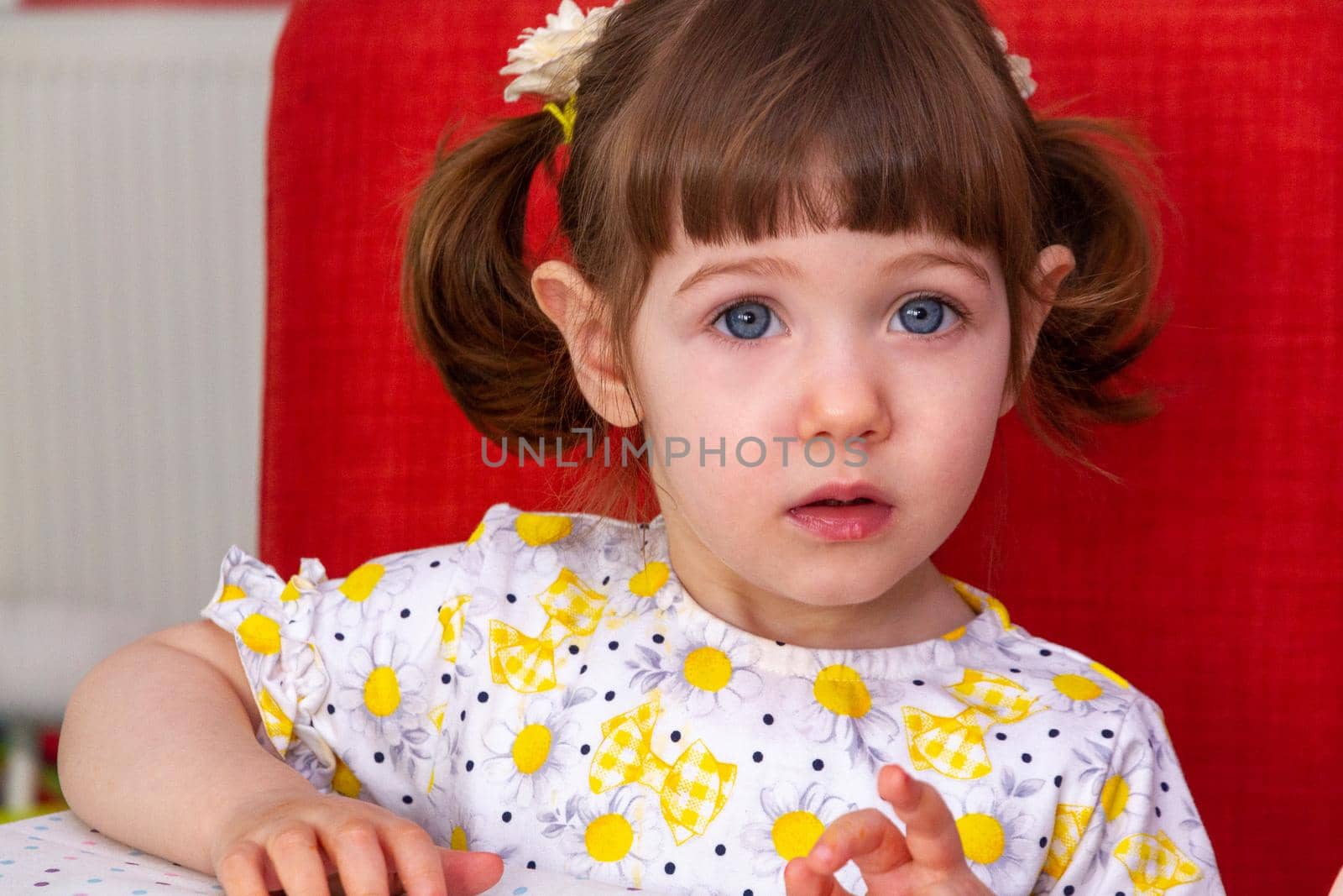 A cute, brown-haired, blue-eyed baby girls with pigtails sitting in an orange chair and looking at camera