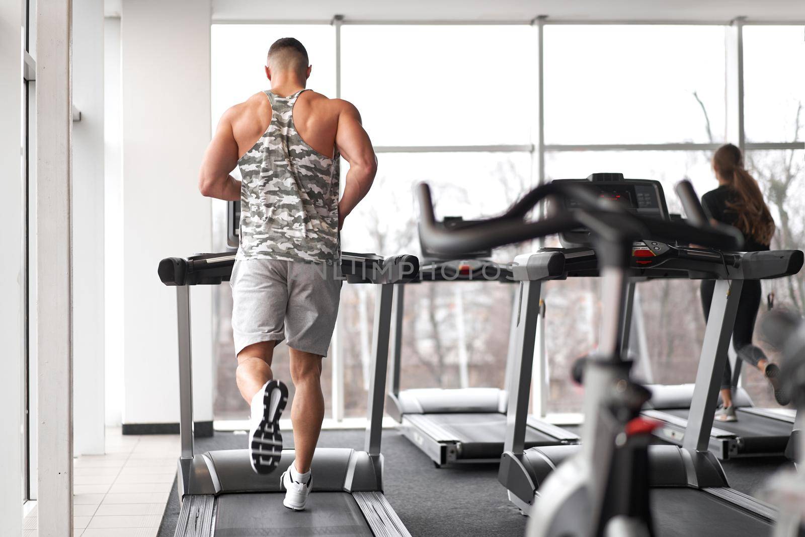 Muscular athletic bodybuilder fitness model running treadmill gym near big window jogging stationary machine do cardio training Weight loss Healthy lifestyle concept. Back view rear full length