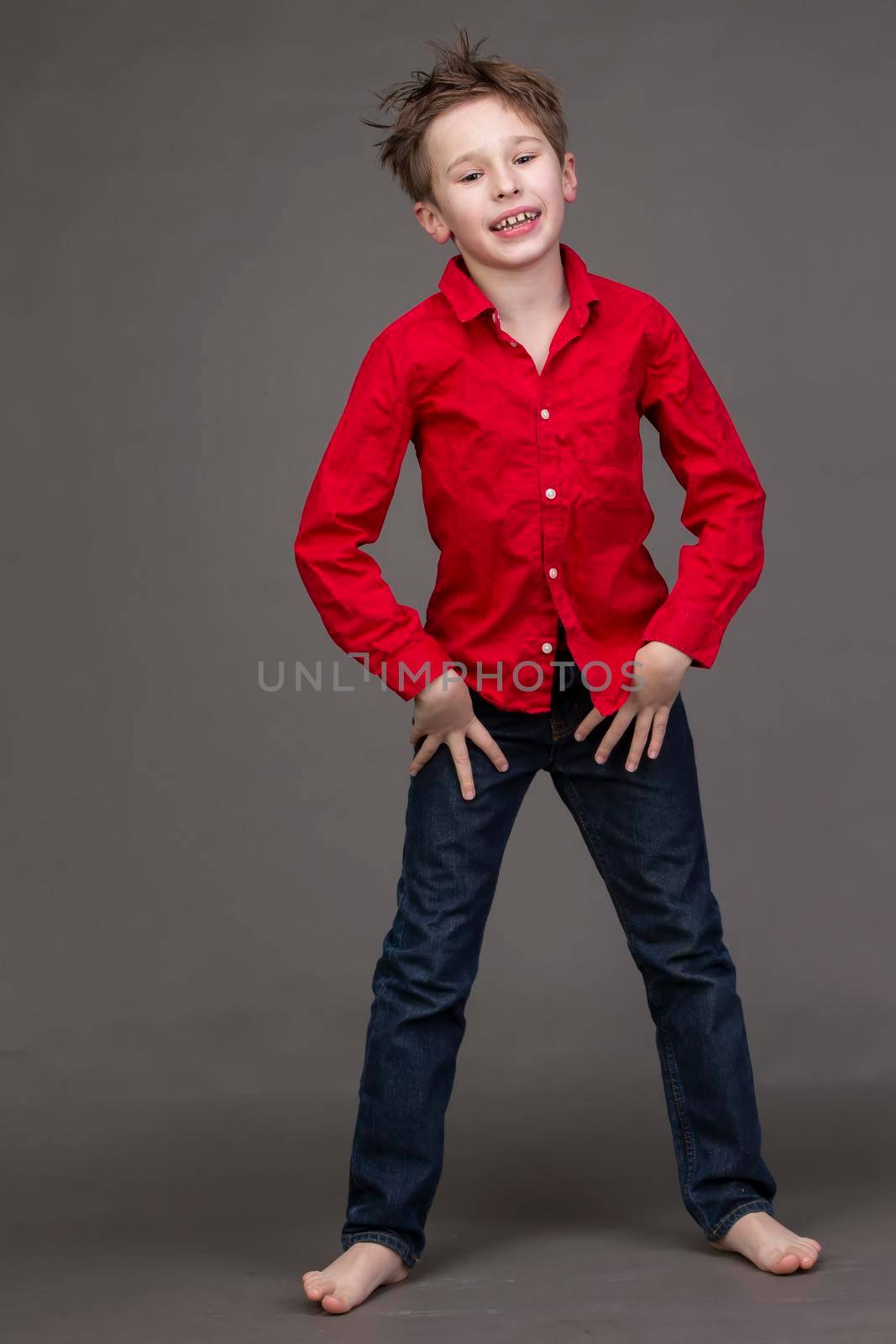 Handsome boy in a red shirt and jeans on a gray background. A child in the modeling business is posing.