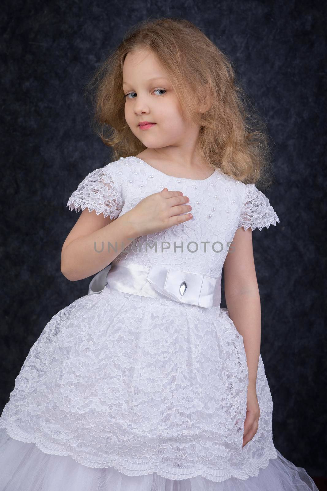 Cute little blonde girl in a beautiful white dress on a dark background. Six year old beautiful girl