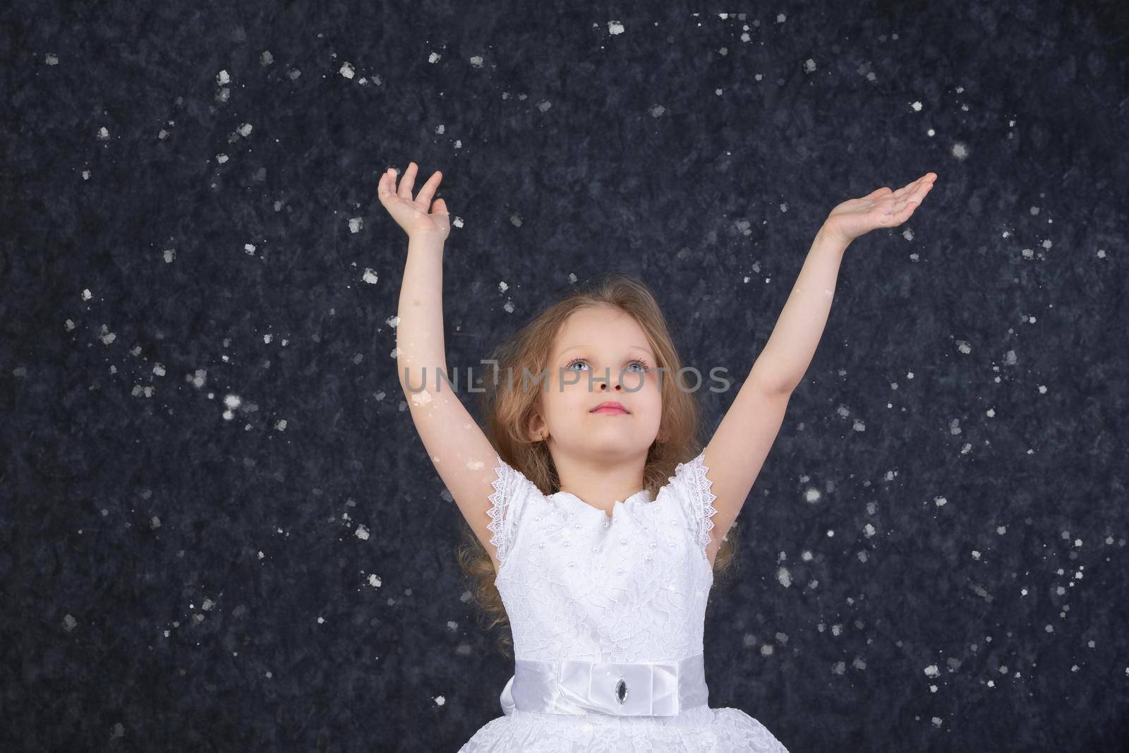 Beautiful little girl in a white dress with raised arms under flying snowflakes.