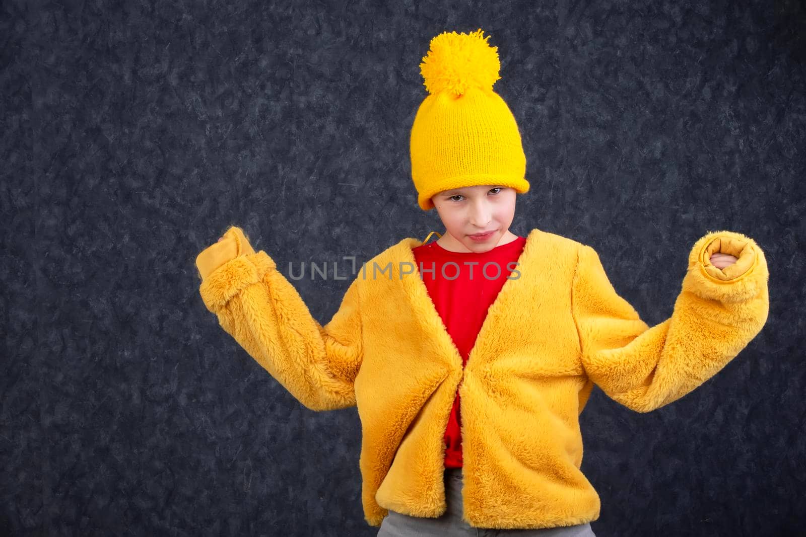 Funny little boy in a fluffy yellow fur coat and a knitted hat on a gray background.