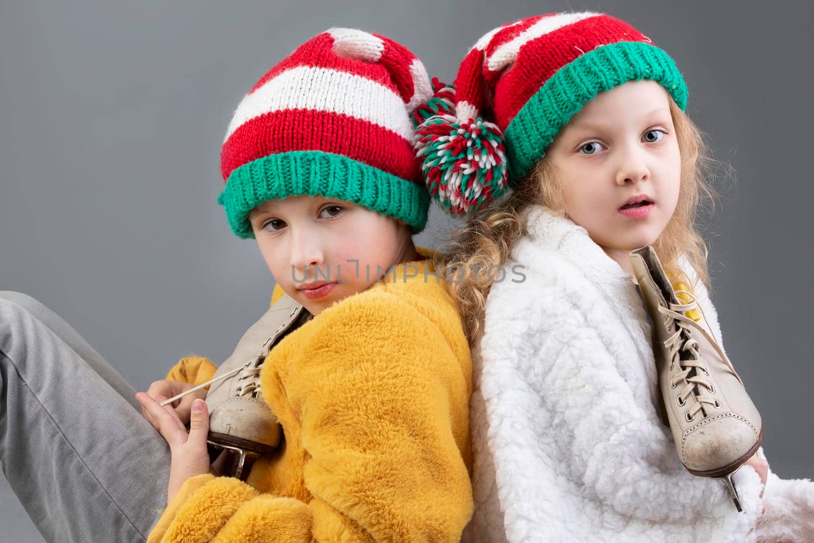 Beautiful little girl and boy in knitted Christmas hats are sitting with their backs to each other and smiling on a gray background. Happy Christmas kids.