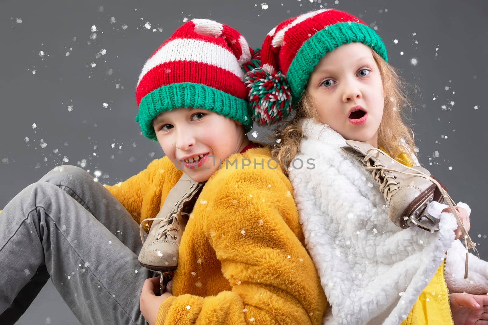 Funny little girl and boy in knitted Christmas hats and vintage ice skates sit with their backs to each other and look at the falling snow on a gray background. Happy Christmas kids.