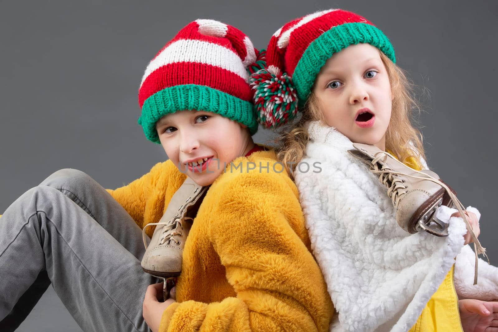 Funny little girl and boy in knitted Christmas hats and vintage ice skates sit with their backs to each other on a gray background. Happy Christmas kids.