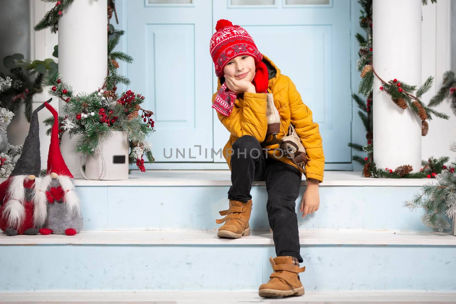 Handsome little boy in christmas. Funny child holds old skates and sits on the porch of a house decorated with Christmas tree decorations