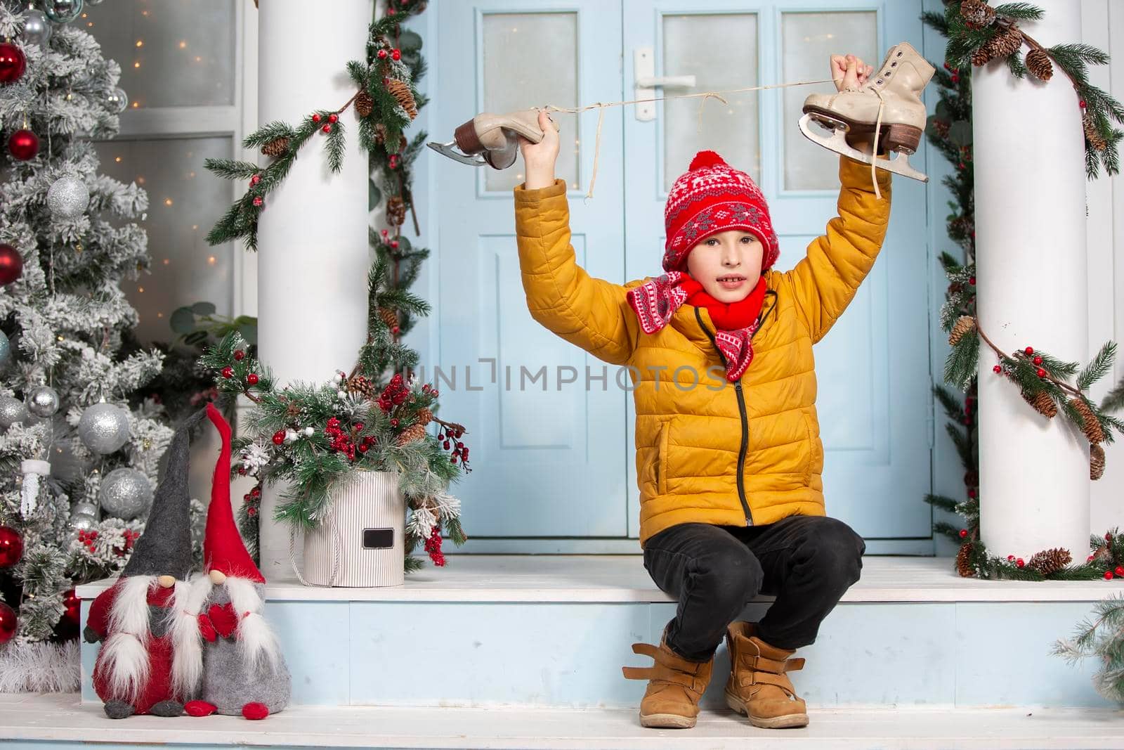 Handsome little boy in christmas. Funny child holds old skates and sits on the porch of a house decorated with Christmas tree decorations
