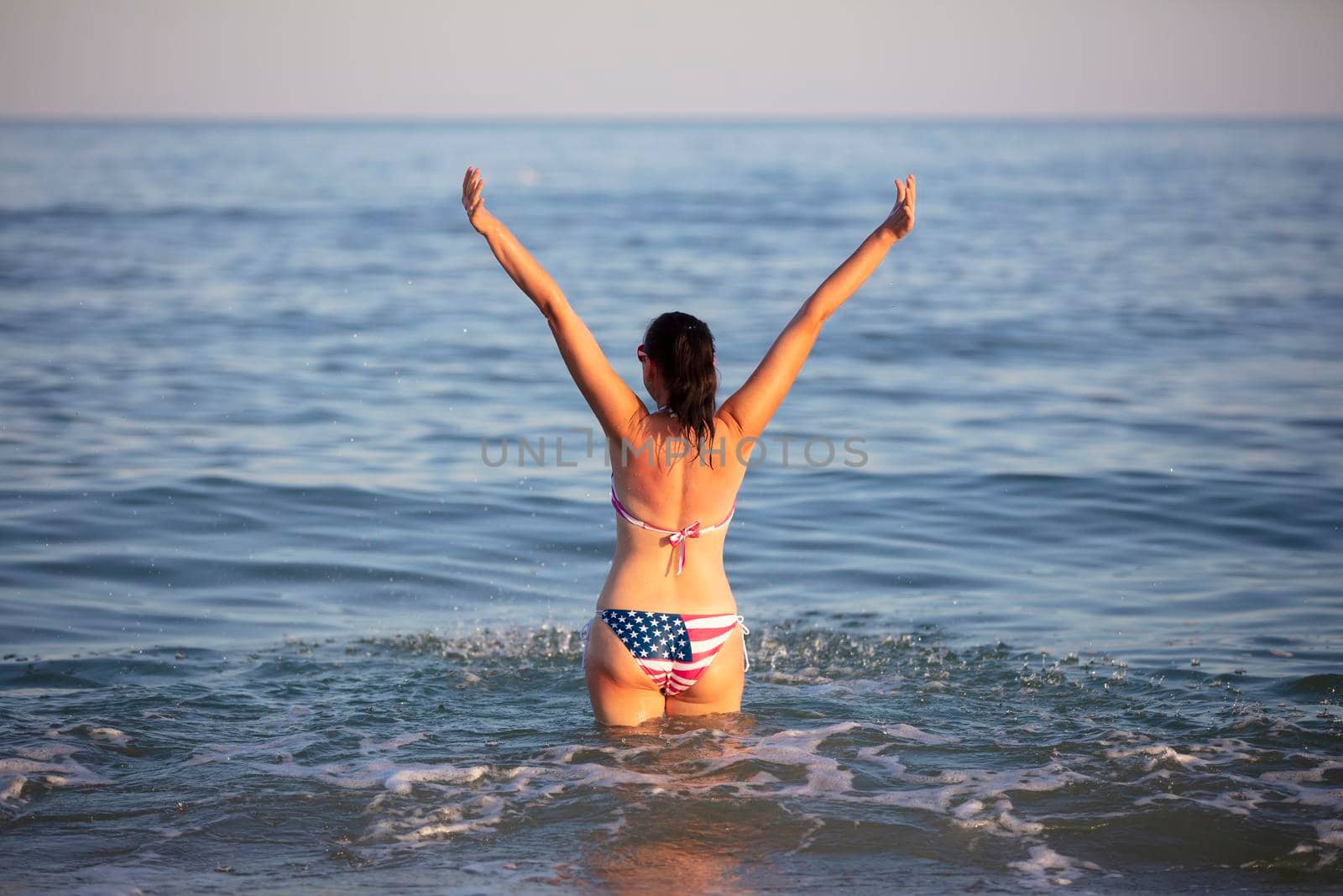A middle-aged woman stands in sea water with her arms raised in a swimsuit with an American flag print.