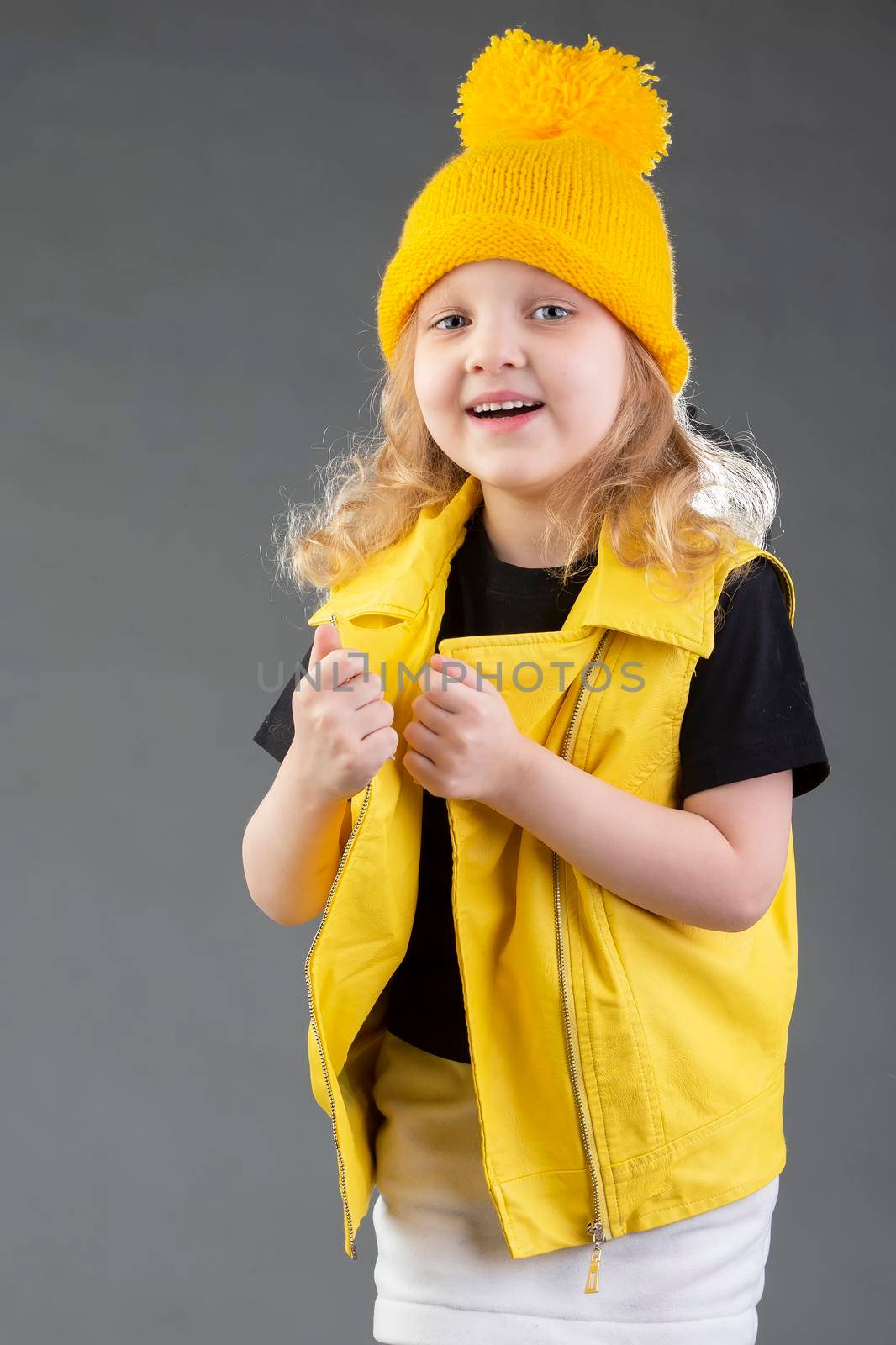Beautiful little blonde girl in a yellow knitted hat on a gray background. Five-year-old cheerful girl.