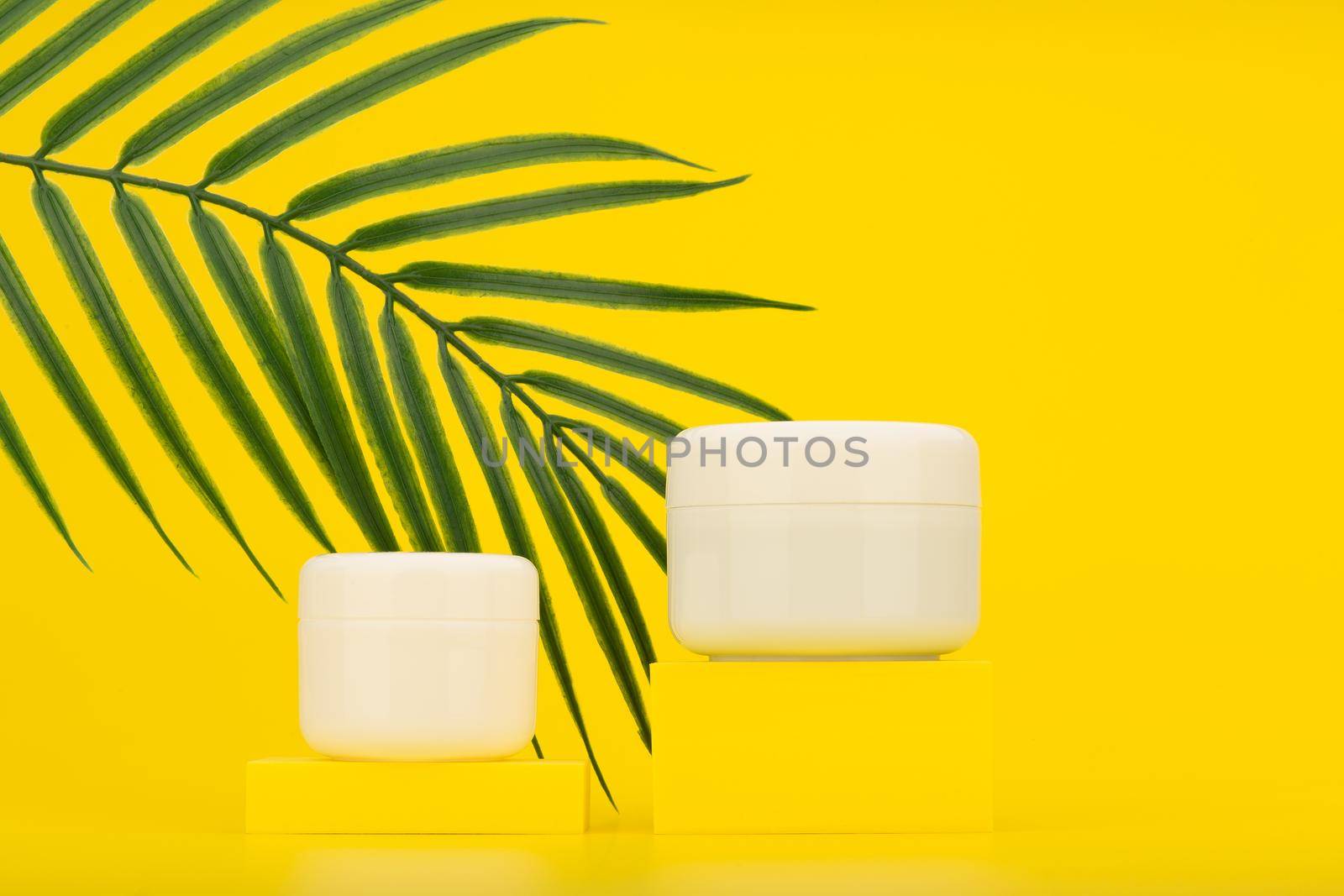 Two cream jars on yellow pedestals against yellow background with palm leaf. Concept of summer skincare with spf by Senorina_Irina