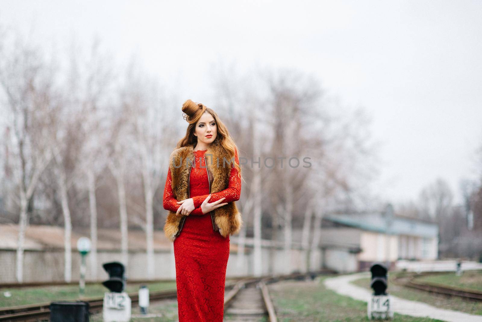 young girl with red hair in a bright red dress on the railway tracks of the station