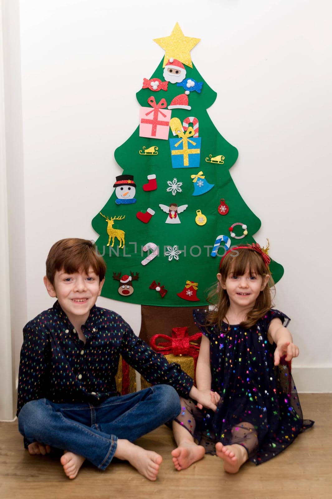 A cute, brown-haired, blue-eyed baby girl and a cute, red-haired, blue-eyed boy smartly dressed sitting in the floor in front of a Christmas tree