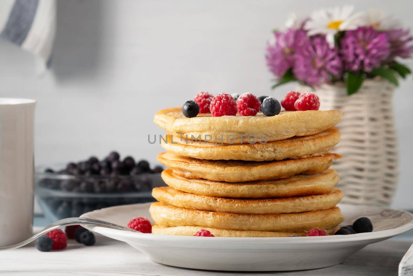 Stack of pancakes with fresh berries on a white plate and a vase of wildflowers on the table.