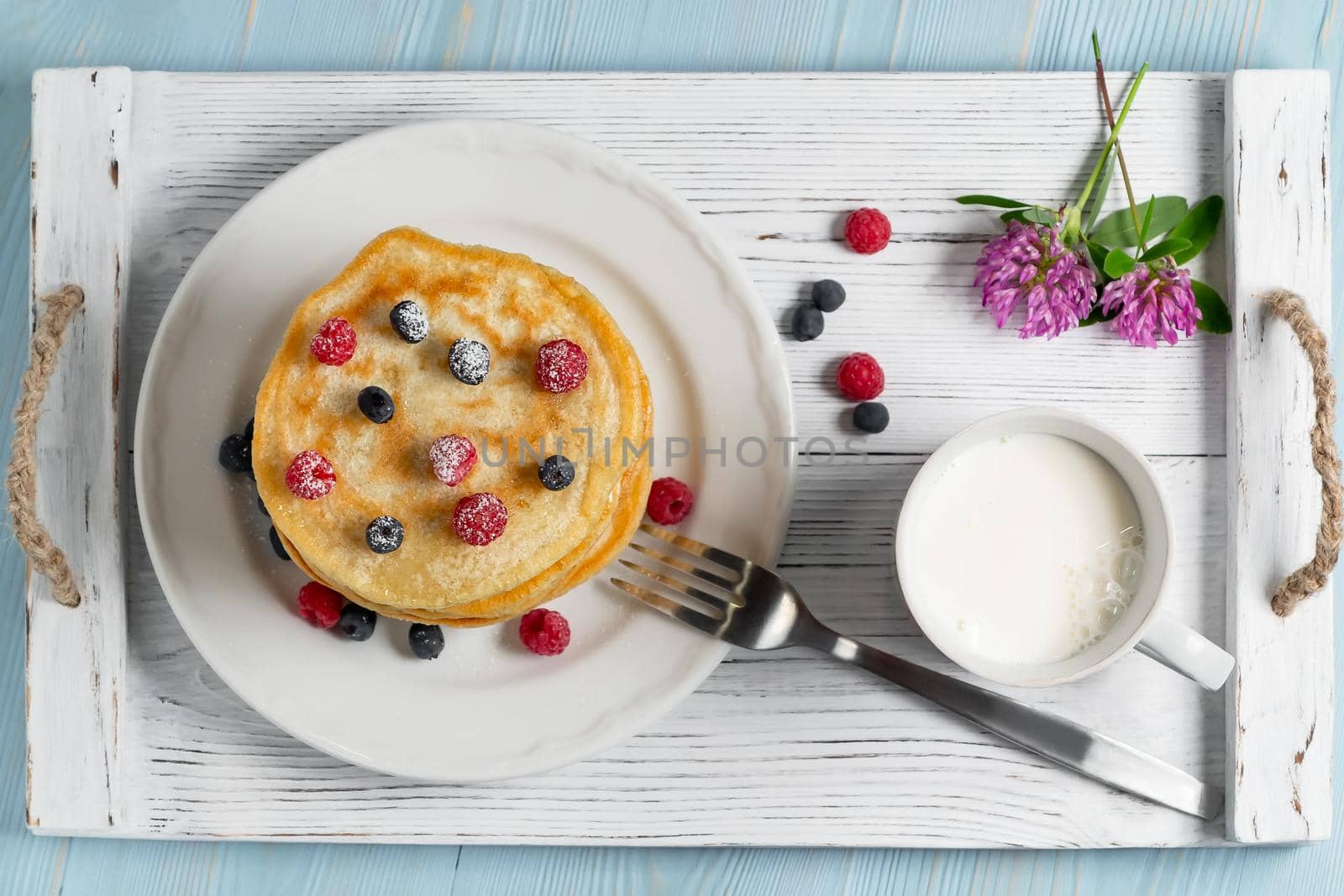 Stack of pancakes with fresh berries on a white plate a cup of milk and wildflowers on the table, top view by galsand