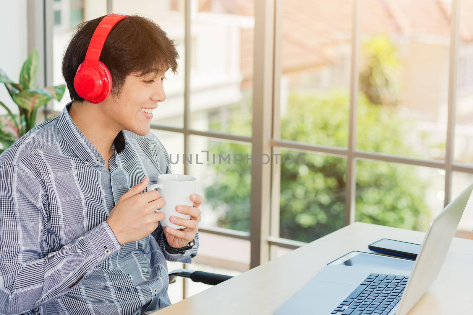 Asian young businessman happiness with red headphones sitting on desk workplace home office with a laptop computer, confident handsome man lifestyle smile relax holding a coffee cup, listens to music