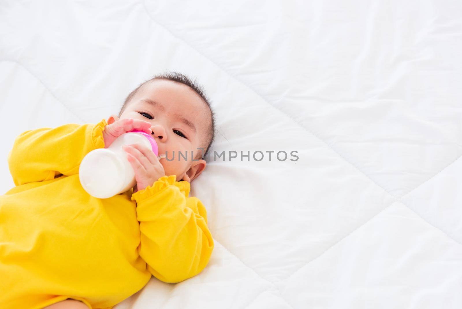 Asian beautiful little baby girl wearing a yellow dress eat milk sleeping feeding lying on the white bed, infant holding a bottle of milk, baby food concept