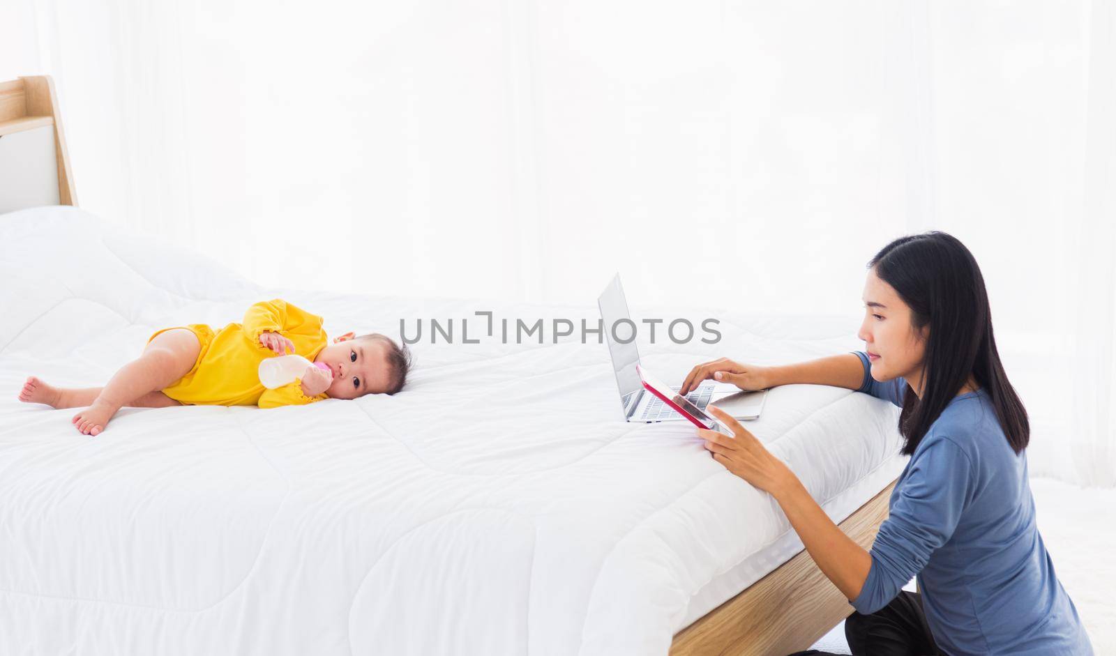 Asian young woman is working busy on laptop computer and tablet at home office in bedroom while her little baby is sleeping feeding lying on the white bed lying near mom