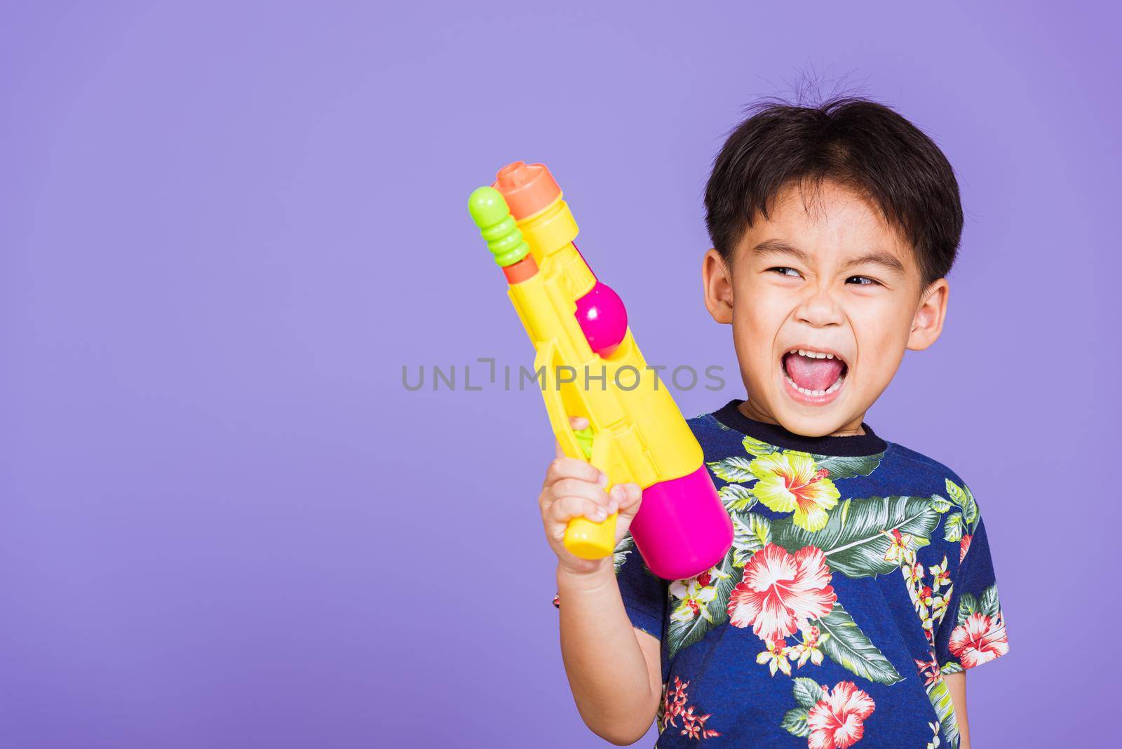 Happy Asian little boy holding plastic water gun, Thai kid funny hold toy water pistol and smiling, studio shot isolated on purple background, Thailand Songkran festival day national culture party