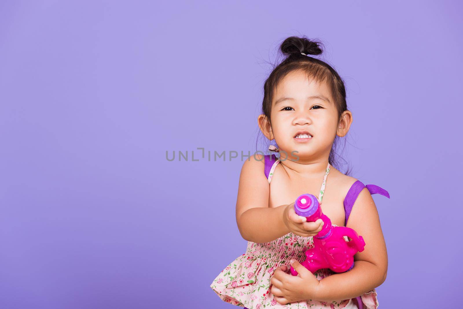 Thai child funny hold toy water pistol and smile, Happy Asian little girl holding plastic water gun, studio shot isolated on purple background, Thailand Songkran festival day national culture concept