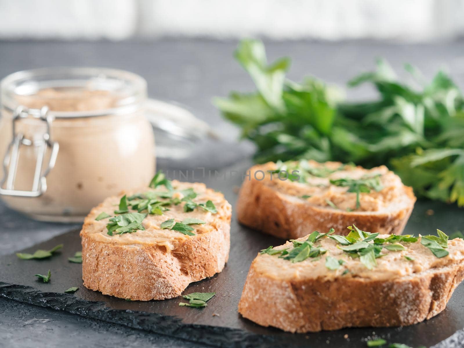 Close up view of slice bread with homemade turkey pate and fresh green parsley on black slate board over gray concrete background, Copy space. Shallow DOF