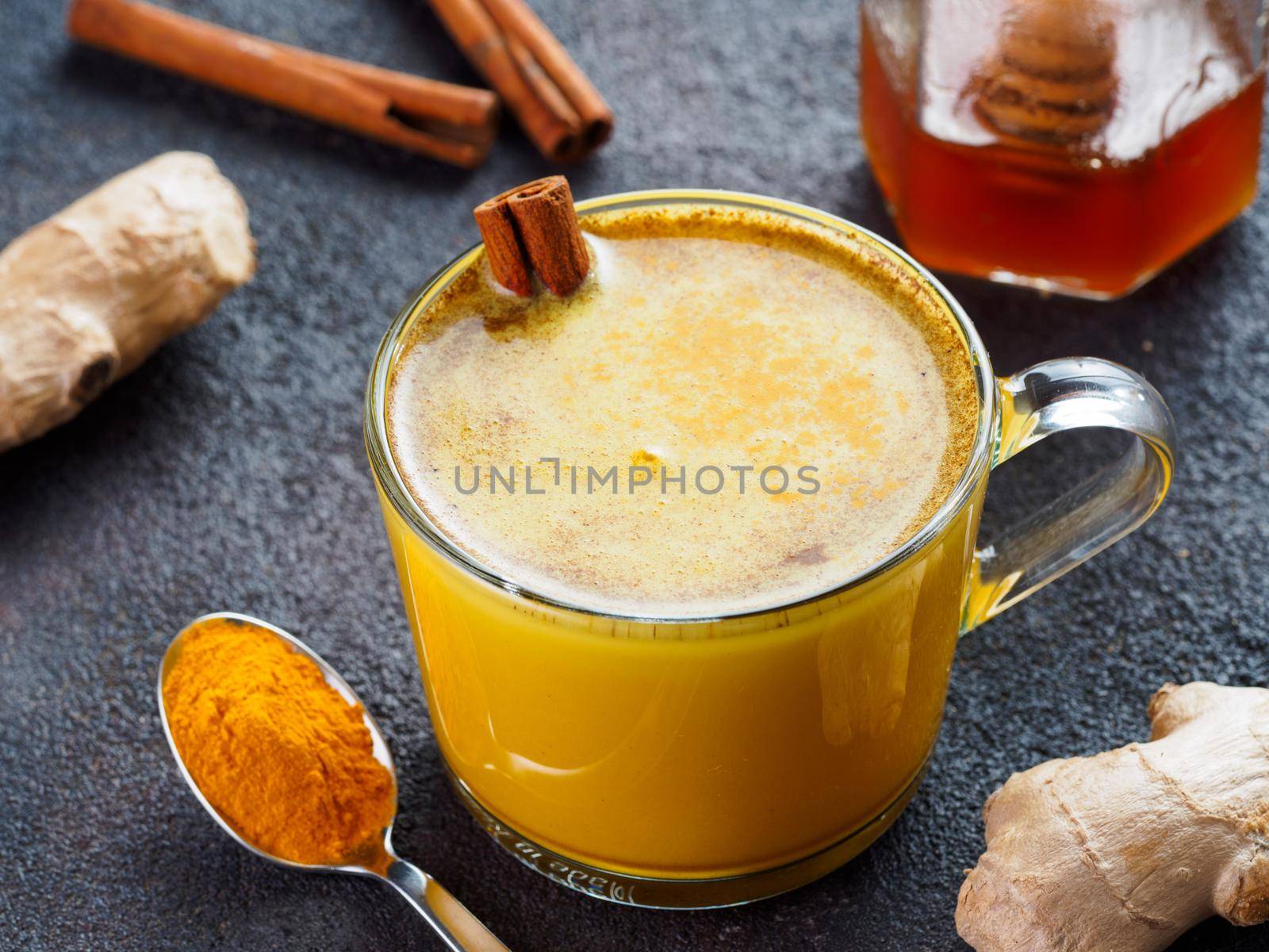 Healthy drink golden turmeric latte in glass cup.Gold milk with turmeric,ginger root,cinnamon sticks,dried turmeric powder and honey over black cement background.Detox turmeric tea and ingredients.