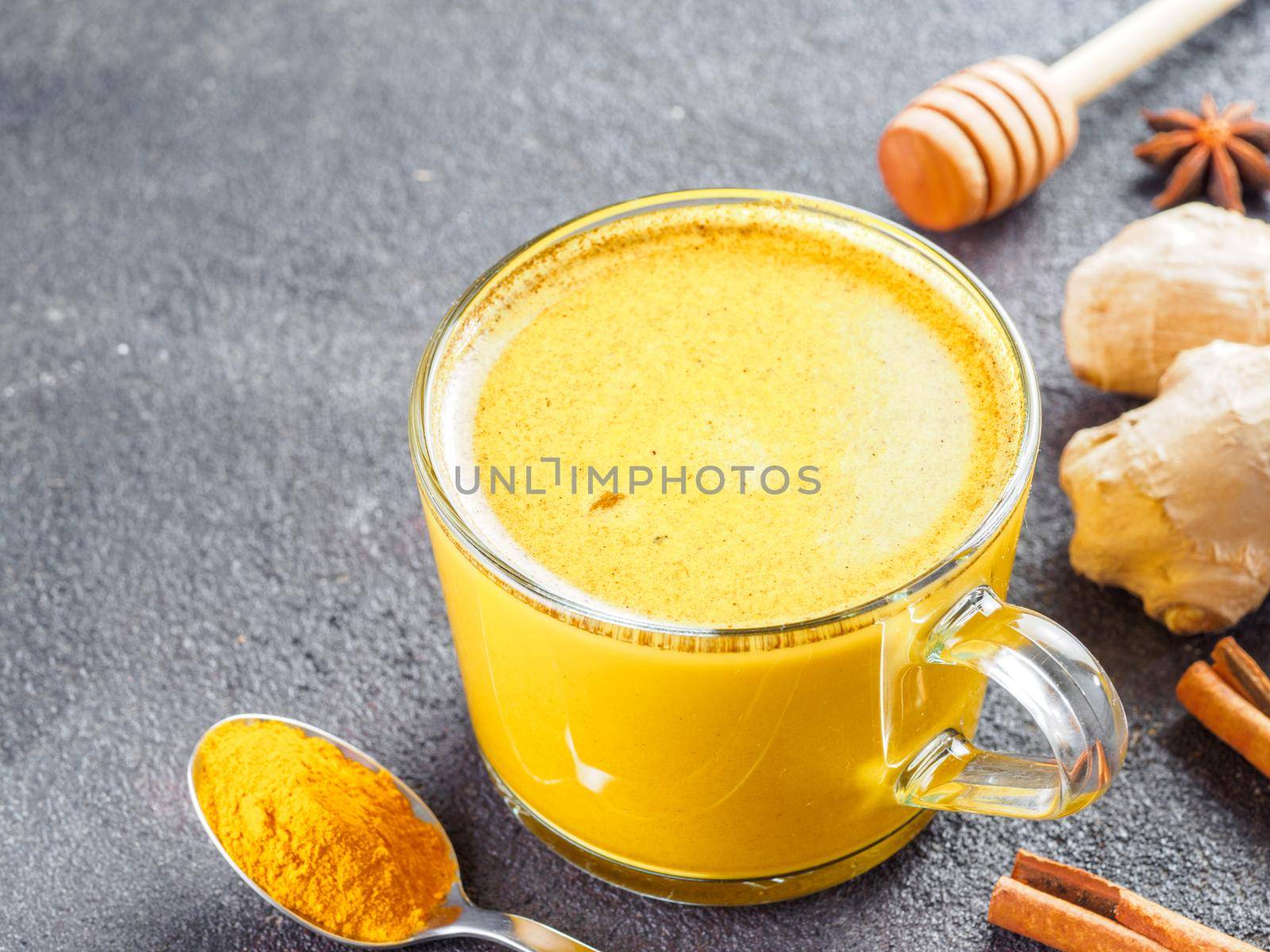 Healthy drink golden turmeric latte in glass cup.Gold milk with turmeric,ginger root,cinnamon sticks,turmeric powder and honey over black cement background.Detox turmeric tea and ingredients.Copyspace
