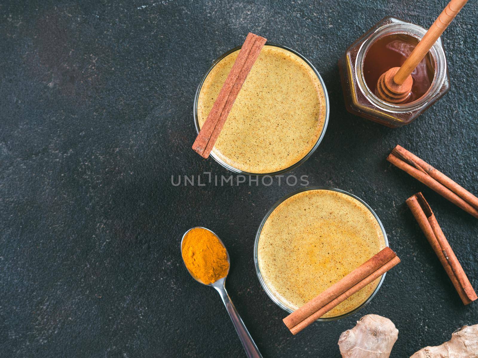Healthy drink golden turmeric latte in glass. Gold milk,ginger root,cinnamon sticks,turmeric powder and honey on black cement background.Detox turmeric tea and ingredients. Copy space. Top view. Toned