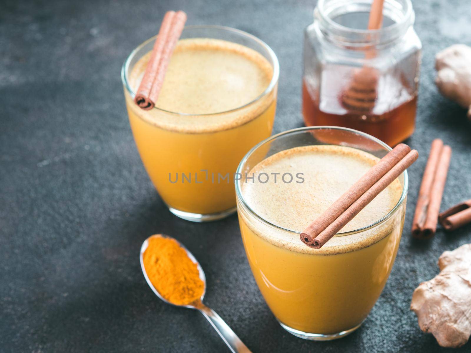 Healthy drink golden turmeric latte in glass.Gold milk with turmeric,ginger root,cinnamon sticks,turmeric powder and honey over black cement background.Detox turmeric tea,ingredients.Copy space.Toned