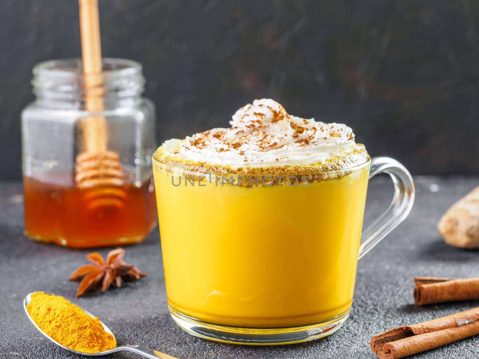 Healthy drink golden turmeric latte with whipped cream in glass cup. Gold milk,ginger root,cinnamon sticks,turmeric powder and honey over black background.Detox turmeric tea and ingredients.Copyspace