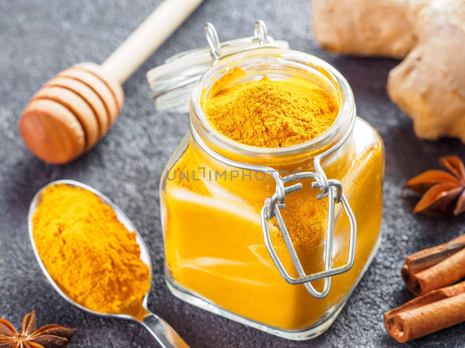 Dried turmeric powder in glass jar and spoon on black cement background. Ingredients for golden milk, turmeric latte or detox drink - honey dipper, ginger root, cinnamon sticks and star anise