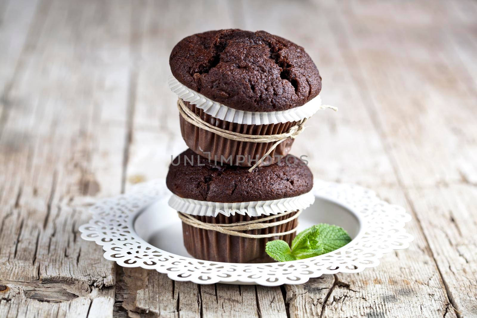 Two fresh dark chocolate muffins with mint leaves on white plate on rustic wooden table by marylooo