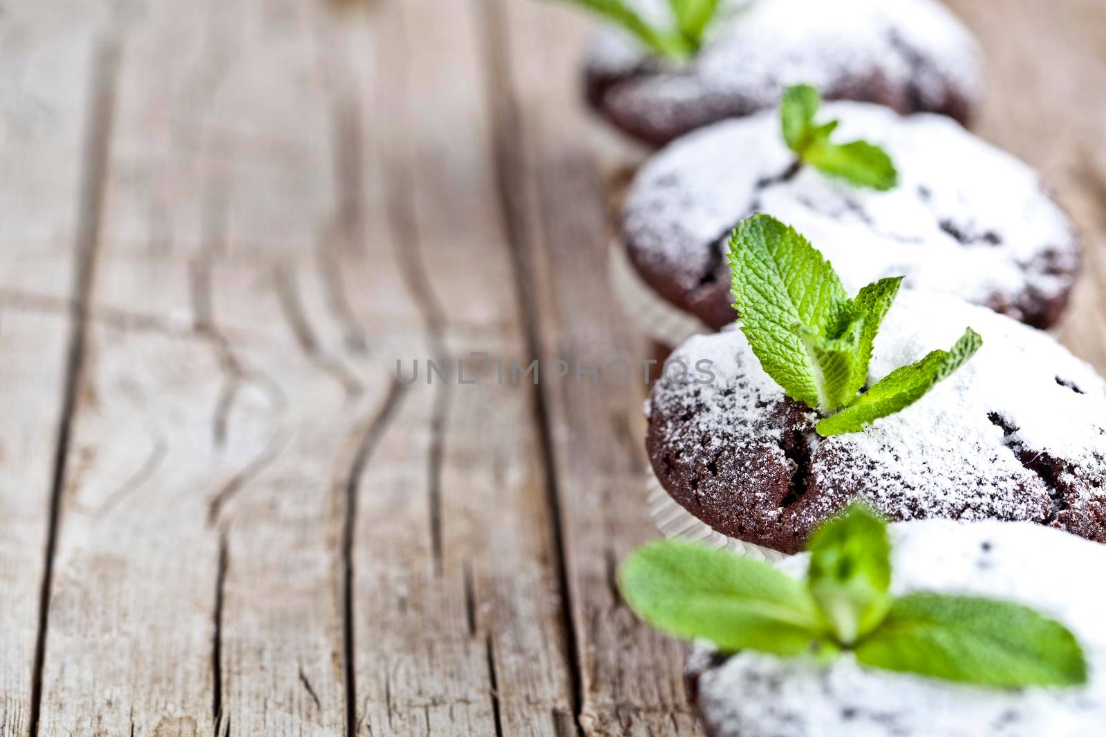 Fresh chocolate dark muffins with sugar powder and mint leaf on rustic wooden table background.  by marylooo
