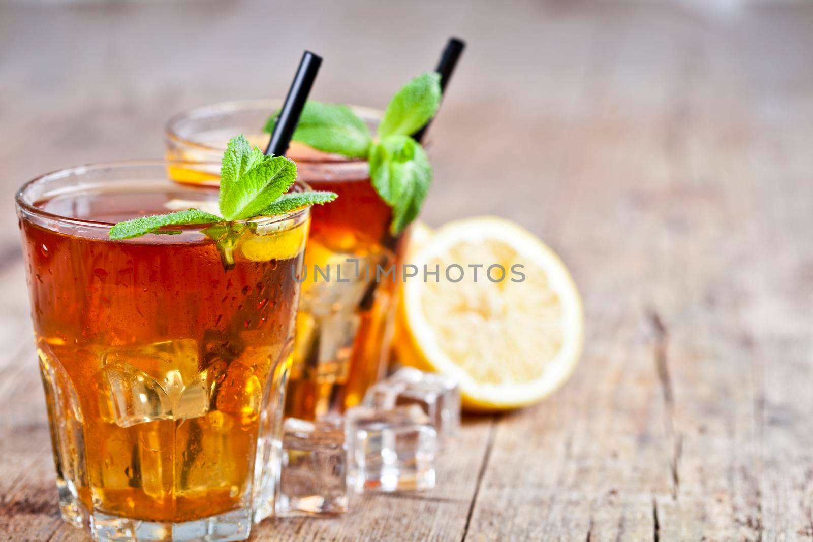 Cold iced tea with lemon, mint leaves and ice cubes in two glasses on rustic wooden table background.  by marylooo