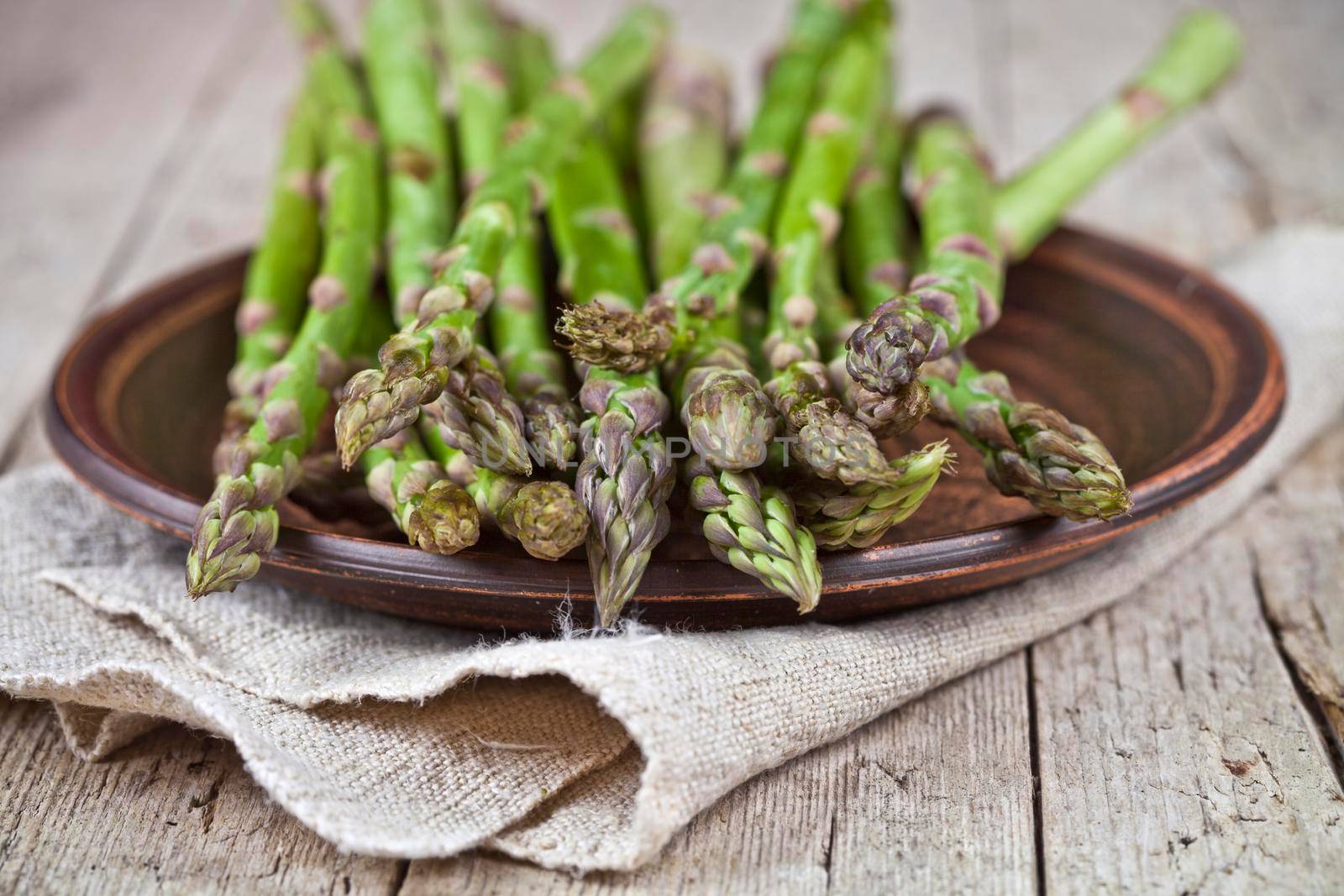 Fresh raw garden asparagus closeup on brown ceramic plate and linen napkin on rustic wooden table background. by marylooo