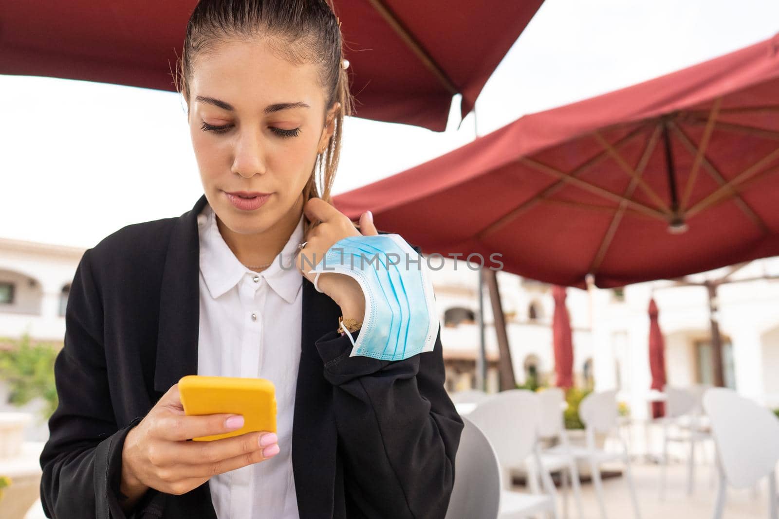 Young millennial woman texting on smartphone at bar table in the city with protective mask in the arm. New normal mobile connections allow to stay in touch with everyone in everywhere
