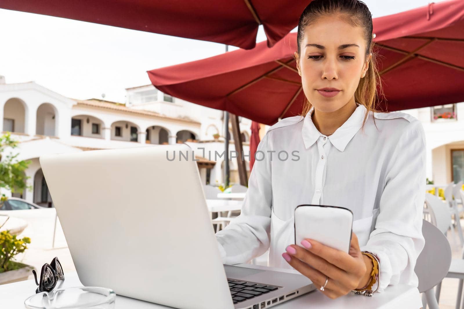 Modern millennial hipster woman using smartphone and laptop at bar table in the city. Internet job choice concept: a young self entrepreneur works on her notebook at a town cafe. New normal concept by robbyfontanesi
