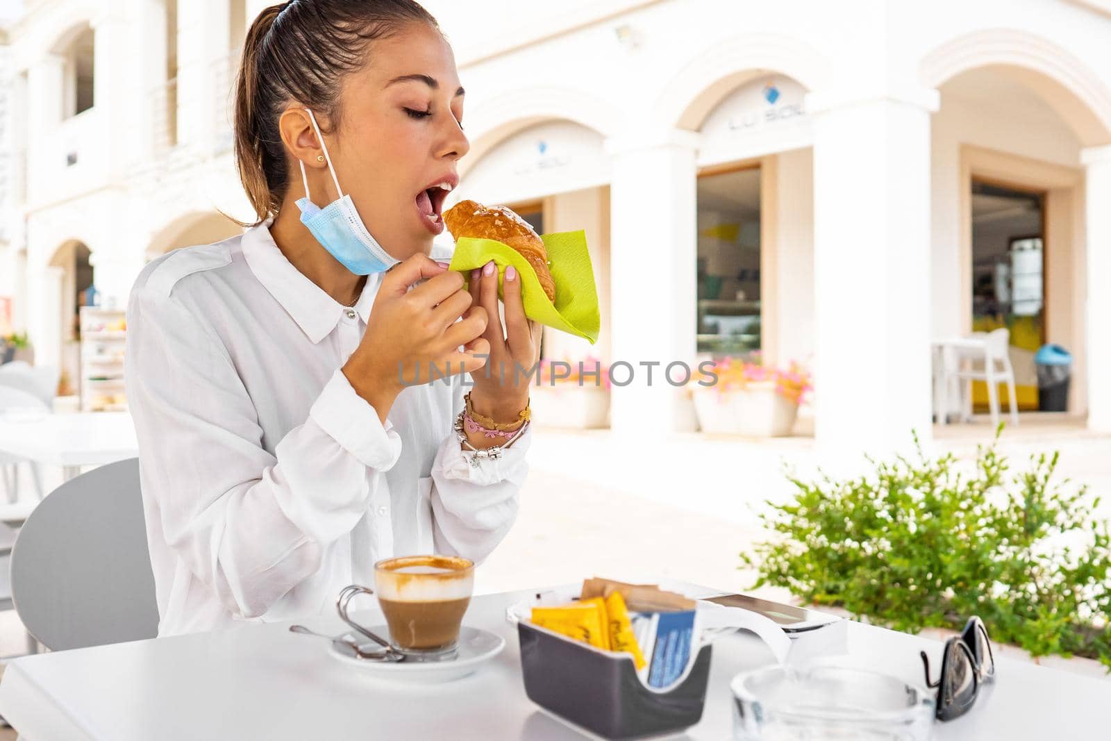 Young woman has breakfast before works at the bar eating a croissant and drinking a cappuccino with the medical protective mask down. New normal city human habits due the Coronavirus pandemic by robbyfontanesi