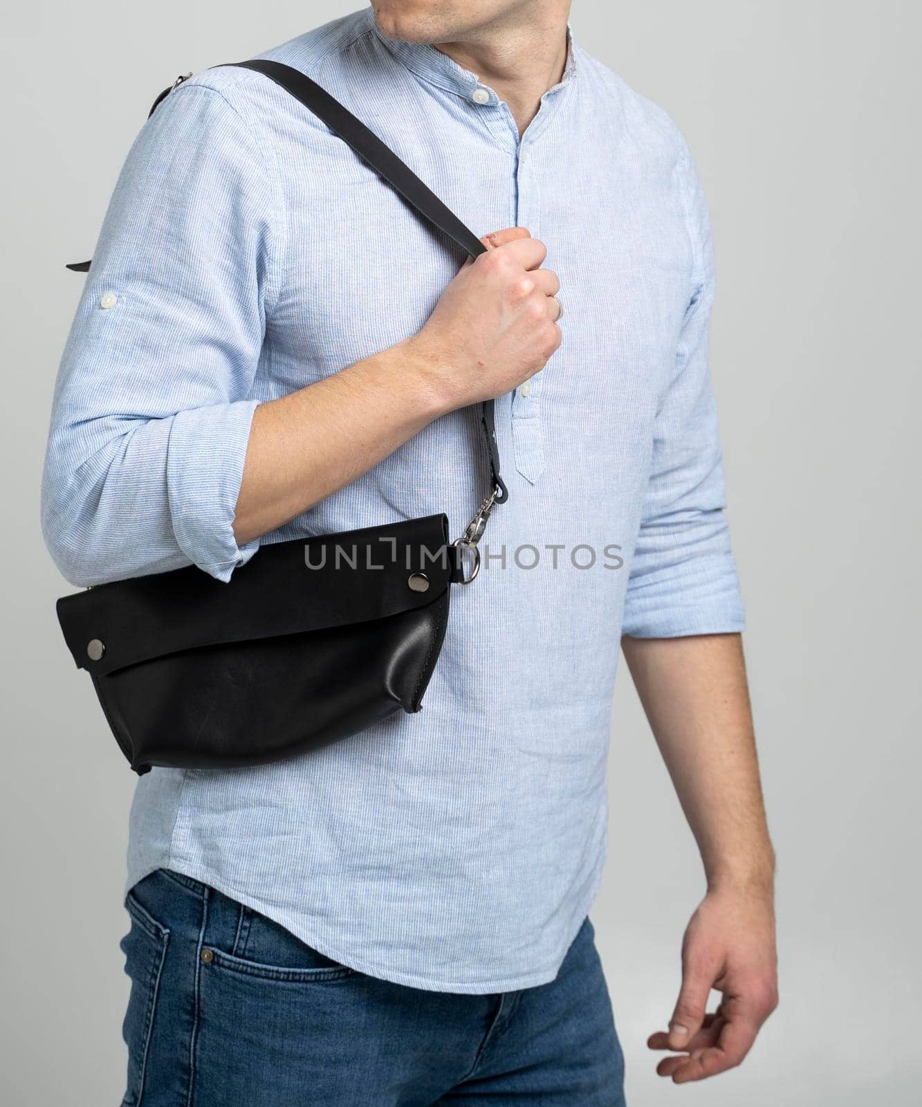 Man in a blue shirt with a leather black handmade bag over his shoulder. Dark designer banana bag. Man in a studio. Comfortable small bag for walking
